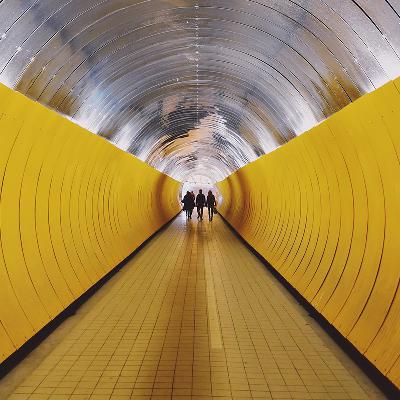 Das Bild hat Symbolcharakter. Drei Personen laufen nebeneinander in einem gelben Tunnel. Sie haben sich wegen gemeinsamer Herausforderungen in der hi!stocks App zusammengeschlossen und sind  nun als Gruppe unterwegs.