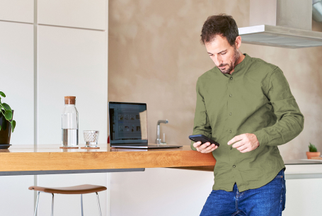 Ein Mann lehnt sich an einen Küchentresen. Über sein Smartphone tätigt er eine Geldanlage. 