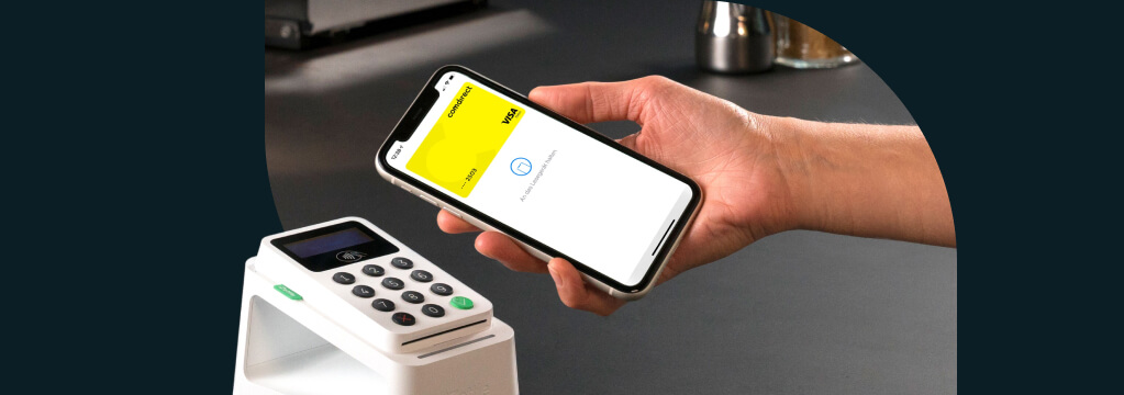 Apple Pay: Eine Hand hält ein Smartphone mit der gelben Visa-Debitkarte an ein Bezahl-Terminal.