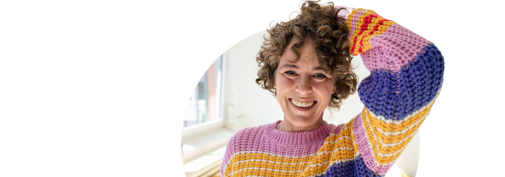 Ein lachende Frau mit einem bunt gestreiftem Strickpulli fasst mit ihrer linken Hand einige Locken am Hinterkopf zusammen. Hinter ihr fällt Licht durch das Fenster einer Altbauwohnung. Die Bauzinsen hat sie passend berechnet. 