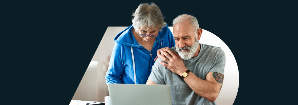 Eine ältere Frau mit Lesebrille steht hinter einem älteren Mann, der an einem Tisch sitzt. Ihre Hände berühren sich an seiner Schulter. Beide schauen gespannt auf das Ergebnis der Immobilienbewertung auf dem Notebook vor ihnen.