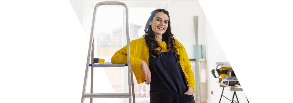 Eine junge Frau steht in einer Wohnung, die gerade renoviert wird. Sie stützt sich auf eine Leiter und lächelt, da sie einen günstigen Modernisierungskredit abschließen konnte.	