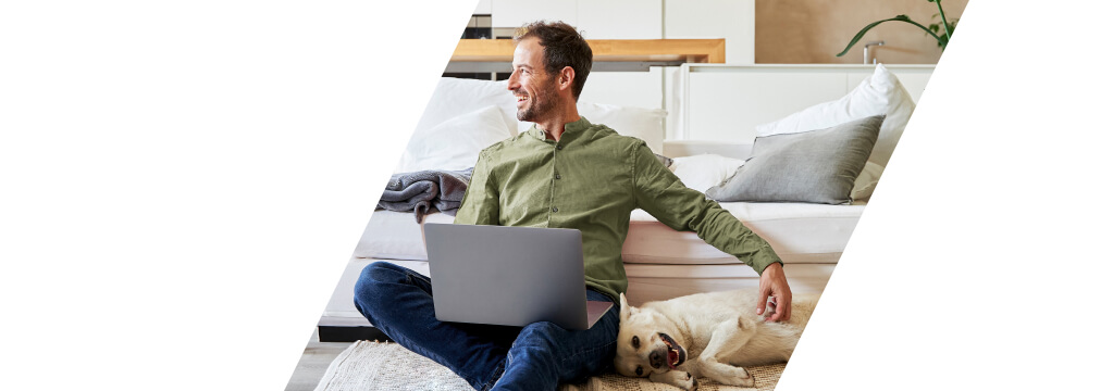 Ein Mann sitzt mit seinem Hund vor einem Sofa und hat ein Notebook auf den Knien liegen. Er freut sich, seine Geldanlage bequem im Blick zu haben. 