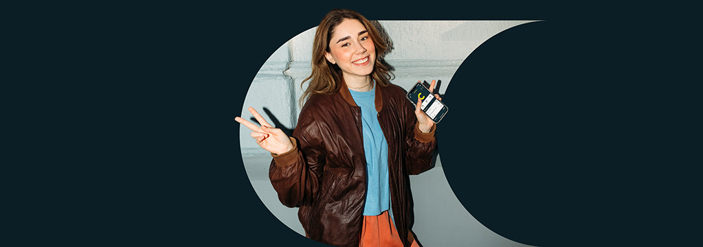 Ein Mädchen in T-Shirt und Bomberjacke lächelt in die Kamera und zeigt dabei den Peace-Zeichen. In der linken Hand hält sie ein Smartphone und öffnet die comdirect-App. Sie freut sich über das Bundle für 18- bis 30-Jährige.