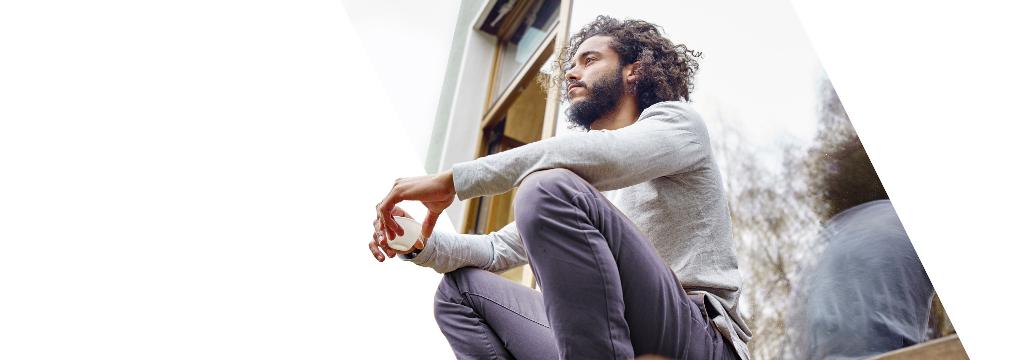 Ein junger Mann mit Bart und Locken sitzt auf einer Fensterbank mit einem Espresso in der Hand und schaut in die Ferne. Er handelt komfortabel mit den Order Funktionalitäten.