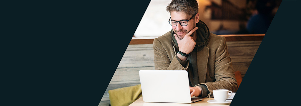 Ein Mann mit gepflegtem Haar, Blazer, modischem Schal, feinem Schnurrbart und Brille sitzt in einem Café und arbeitet am Laptop. Er freut sich auf den neuen ProTrader mit interaktiver Realtime-Handelsoberfläche.