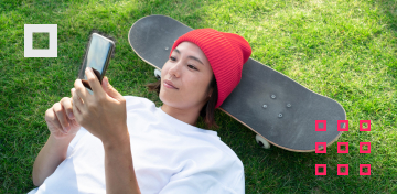 Eine Teenagerin mit roter Mütze liegt mit dem Kopf auf einem Skateboard und schaut auf ihr Smartphone-Display.