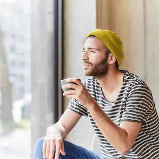 Ein Mann mit gelber Mütze sitzt entspannt mit einer Kaffeetasse vor einem bodentiefen Fenster und schaut nach draußen.