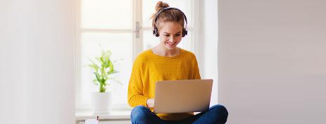 Eine junge Frau mit gelbem Pullover und Headset sitzt im Schneidersitz und balanciert ihren Laptop auf den Knien. Sie nimmt an einem Online-Seminar teil.