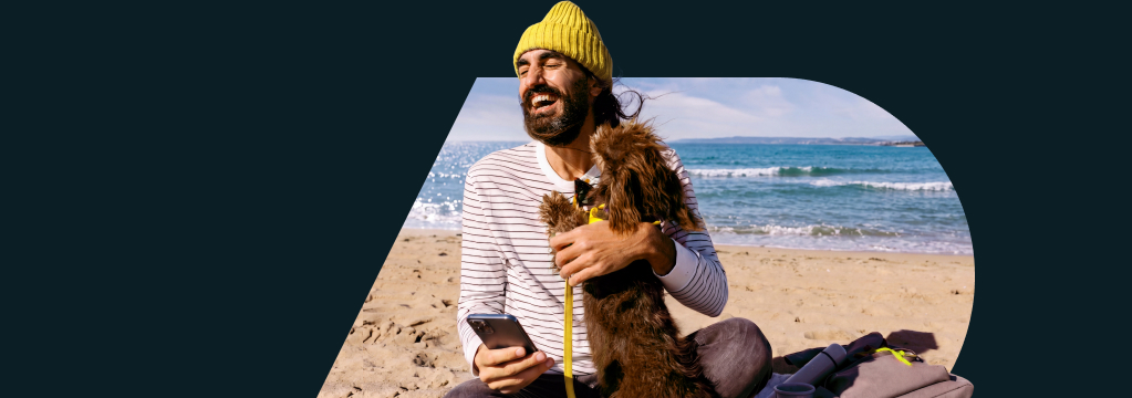 Ein lachender Mann mit gelber M&uuml;tze sitzt am Strand, h&auml;lt ein Smartphone in der Hand und umarmt einen kleinen Hund. Es gibt den Hinweis „Bis zu 150 Euro Pr&auml;mie“.