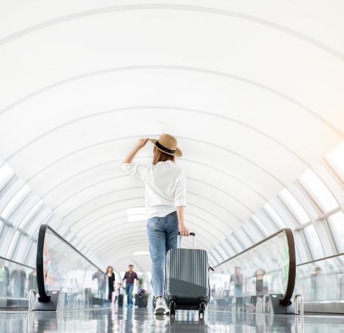 Die Rückenansicht einer Frau mit Strohhut und Rollkoffer am Flughafen.