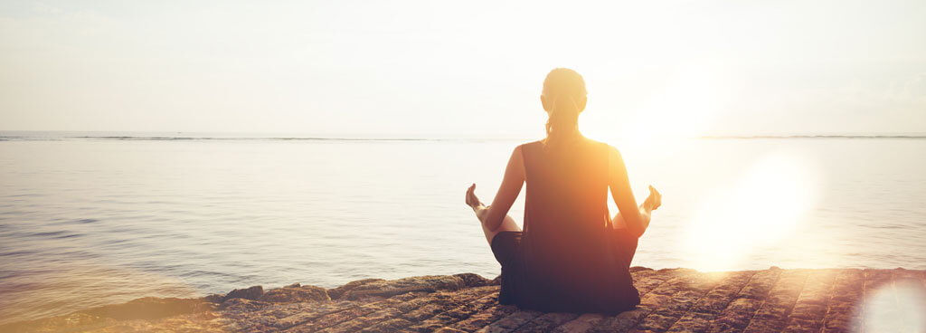 Eine meditierende Frau sitzt am Wasser vor einem Sonnenuntergang und symbolisiert hiermit die Möglichkeit, in den boomenden Gesundheitssektor zu investieren.