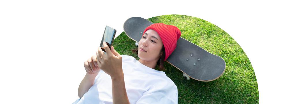 Eine junge Frau mit roter Mütze liegt mit dem Kopf auf einem Skateboard und schaut auf ihr Smartphone-Display. Sie handelt Fonds mit comdirect.