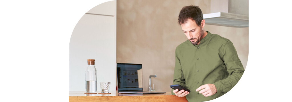 Ein Mann im grünen Hemd schaut konzentriert auf sein Smartphone. Seinen Laptop hat er auf einer Küchentheke abgestellt.