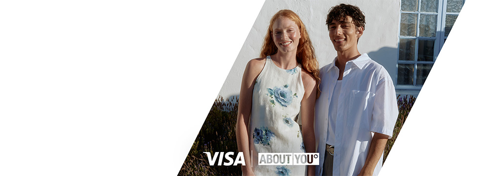 Eine rothaarige Frau im geblümten Sommerkleid und ein gelockter Mann im weißen Hemd lächeln in die Sonne. Am unteren Bildrand ist das weiße Visa-Logo zu sehen.