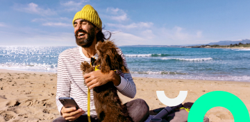 Ein lachender Mann mit gelber M&uuml;tze sitzt am Strand, h&auml;lt ein Smartphone in der Hand und umarmt einen kleinen Hund.