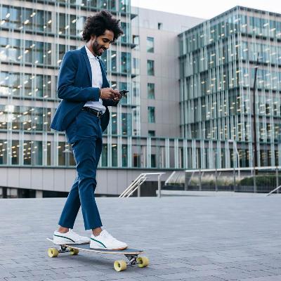 Ein junger Mann im blauen Anzug schaut entspannt auf sein Smartphone. Er rollt auf einem Skateboard vor einem Bürogebäude entlang.