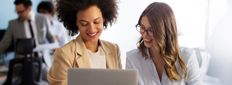 Zwei junge Frauen stehen in einem Büro und schauen gemeinsam auf einen Laptopbildschirm.