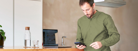 Ein Mann im grünen Hemd schaut konzentriert auf sein Smartphone. Seinen Laptop hat er auf einer Küchentheke abgestellt.