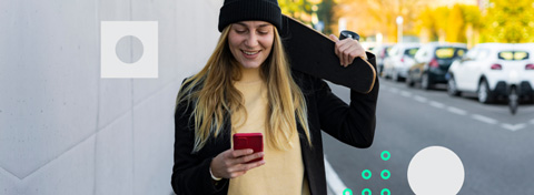 Eine Frau mit einem schwarzen Skateboard auf der Schulter schaut fröhlich auf ihr rotes Smartphone.