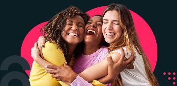 3 junge Frauen sitzen auf einem Sofa, umarmen sich und lachen.
