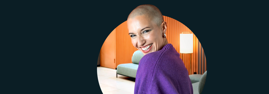 Eine geschminkte Frau mit Glatze und Ohrring in einem violetten Strickpullover steht in einem modern eingerichteten Wohnzimmer und schaut freudig lächelnd über ihre Schulter in die Kamera.