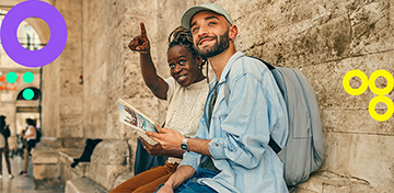 Eine Frau zeigt mit ihrem Finger in die Ferne, während ein neben ihr sitzender Mann mit einem Reiseführer in den Händen ihrem Blick folgt.